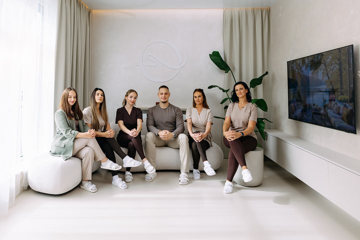 Šest členů týmu Harmony Haven Spa v pracovním oblečení sedí na světlých sedacích prvcích v čekací části s logem na stěně a televizí