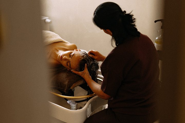 Pracovnice myje vlasy klientce u mycího boxu, pohled škvírou mezi dveřmi